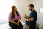 A medical professional takes notes on a clipboard while talking with a patient.