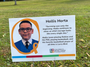 A sign in a grassy field shows a photo of a boy surrounded by a gold border with a gold awareness ribbon for Childhood Cancer Awareness Month. His name, Hollis Horta, appears to the side, along with a quote about his courage and a list of some of his favorite activities. The Children's Brain Tumor Foundation logo appears at the bottom of the sign.