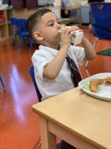 A 4-year-old boy wears a white shirt and tie, which appear to be part of a school uniform, and sits at a small table in a classroom. He's drinking from a small carton of milk, which he's holding with both hands, and has food on a plate in front of him.