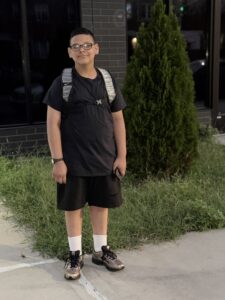A sixth-grade boy stands outside in front of a building and smiles for a photo. He's wearing a black T-shirt and shorts, sneakers, glasses, and his backpack.