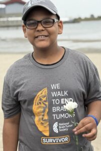 A sixth-grade boy smiles for a photo on the beach while holding a white flower. He's wearing a gray T-shirt and baseball cap, both featuring the National Brain Tumor Society logo. His shirt says "We walk to end brain tumors" and identifies him as a survivor.