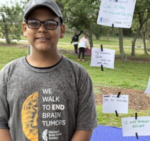 A sixth-grade boy smiles for a photo next to a message he wrote on a small piece of paper, which is displayed in a park next to other messages. His reads "walk to the end for brain tumor" and the paper next to his, written by his mom, reads "I walk for my son Hollis Horta." The boy is wearing a gray National Brain Tumor Society T-shirt that says "we walk to end brain tumors."