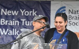 A woman and her son stand side by side before a podium and microphone. Banners hung behind them show that they're at the National Brain Tumor Society's New York Brain Tumor Walk.