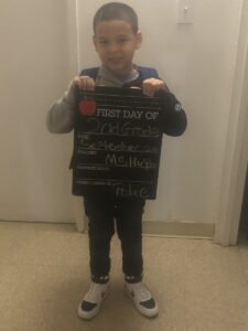 A young boy smiles proudly while holding up a black chalkboard sign that reads "first day of second grade."
