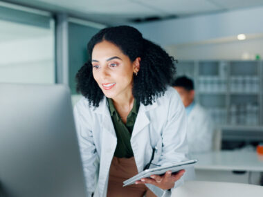A woman in a white coat holds a tablet and looks at a computer screen.